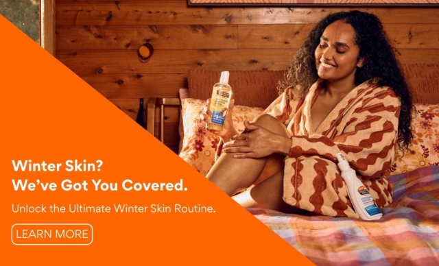 Unlock the Ultimate Winter Skin Routine with Palmer's Cocoa Butter - A smiling women shown applying Palmer's Cocoa Butter Body Oil to legs whilst on a bed in a fluffy robe. 